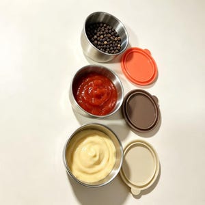 Op de afbeelding: Drie ronde roestvrijstalen bakjes gevuld met voedsel. Het bovenste bakje bevat zwarte peperkorrels, het middelste rode tomatensaus en het onderste een romige gele saus. Elk bakje heeft een bijpassend deksel in oranje, bruin of beige.