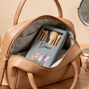 Puede incluir: Un bolso de maquillaje color canela abierto revela un estuche gris para brochas de maquillaje con el nombre "Grace" y detalles florales. El bolso contiene un juego de brochas de maquillaje. Otros artículos de maquillaje, incluyendo base, lápiz labial y un espejo, están sobre la mesa.