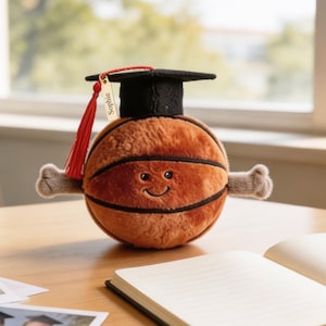 May include: A plush basketball toy with a smiling face, wearing a black graduation cap with a red tassel. A small rectangular gold-colored tag hangs from the tassel, featuring the name Sophie engraved in black text on its surface.