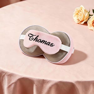 K&ouml;nnte beinhalten: Eine rosa Schlafmaske mit dem Namen "Thomas" in schwarzer Schrift. Die Maske hat ein wei&szlig;es elastisches Band und eine graue Innenseite. Zwei pfirsichfarbene Rosen liegen auf einer rosa Oberfl&auml;che.