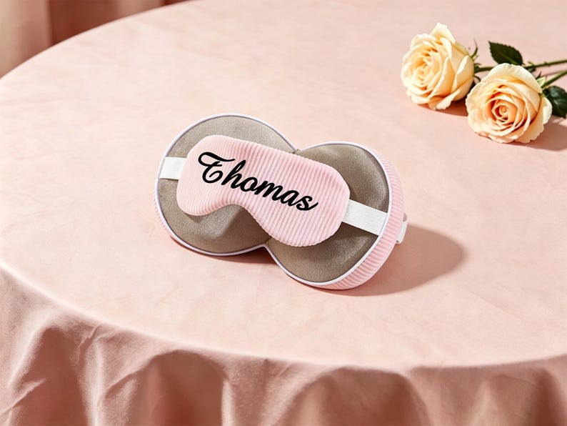 K&ouml;nnte beinhalten: Eine rosa Schlafmaske mit dem Namen "Thomas" in schwarzer Schrift. Die Maske hat ein wei&szlig;es elastisches Band und eine graue Innenseite. Zwei pfirsichfarbene Rosen liegen auf einer rosa Oberfl&auml;che.