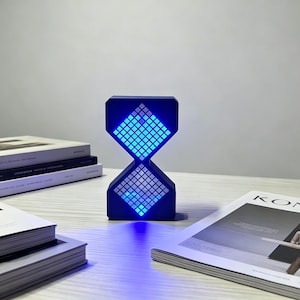 May include: A blue, hourglass-shaped digital clock with a grid of blue LED lights. The clock sits on a light surface, with books and a magazine. The clock's design is modern and minimalist, suitable for a desk or bedside table.