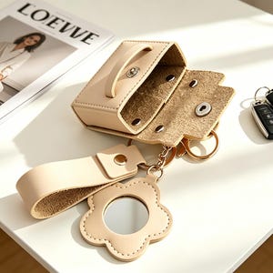 May include: A beige leather key pouch with a snap closure, featuring a matching wrist strap and a flower-shaped charm with a small circular mirror. The pouch is open, revealing a textured interior, and is attached to a metal key ring.