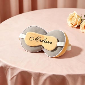 K&ouml;nnte beinhalten: Eine graue und gelbe Schlafmaske mit dem Namen "Madison" in Schreibschrift. Die Maske hat ein wei&szlig;es Gummiband und liegt auf einer rosa Tischdecke. Zwei pfirsichfarbene Rosen sind im Hintergrund.