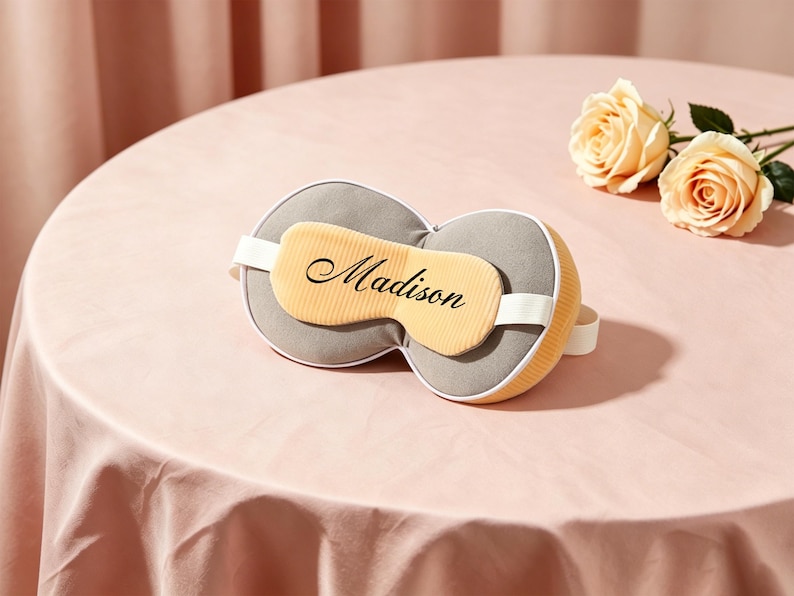 K&ouml;nnte beinhalten: Eine graue und gelbe Schlafmaske mit dem Namen "Madison" in Schreibschrift. Die Maske hat ein wei&szlig;es Gummiband und liegt auf einer rosa Tischdecke. Zwei pfirsichfarbene Rosen sind im Hintergrund.