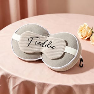 K&ouml;nnte beinhalten: Ein graues Reisekissen- und Augenmasken-Set mit dem Namen "Freddie" in Schreibschrift. Das Set enth&auml;lt eine Rei&szlig;verschlusstasche, einen wei&szlig;en Riemen und liegt auf einer rosa Oberfl&auml;che mit zwei Pfirsichrosen.