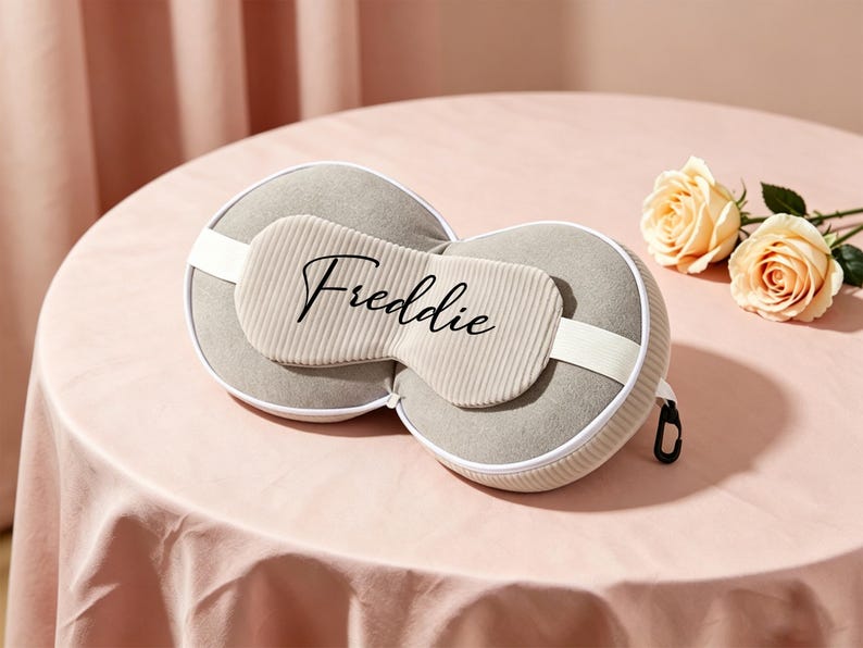 K&ouml;nnte beinhalten: Ein graues Reisekissen- und Augenmasken-Set mit dem Namen "Freddie" in Schreibschrift. Das Set enth&auml;lt eine Rei&szlig;verschlusstasche, einen wei&szlig;en Riemen und liegt auf einer rosa Oberfl&auml;che mit zwei Pfirsichrosen.