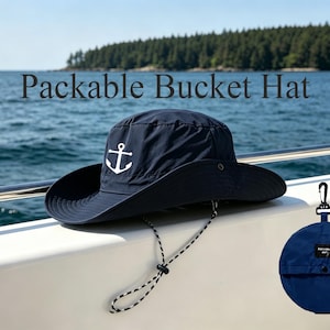Peut inclure: Un chapeau bob bleu marine avec un motif d'ancre blanc sur le devant et une mentonnière réglable noire et blanche. Le texte Packable Bucket Hat est visible au-dessus. Une version pliée du chapeau avec un clip noir est également présente.