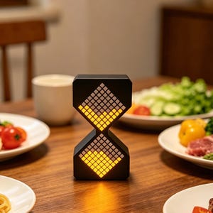 May include: A black hourglass-shaped lamp with a pixelated design, emitting a warm yellow glow. The lamp sits on a wooden table, surrounded by plates of food, including vegetables, pasta, and steak. The background is softly blurred.