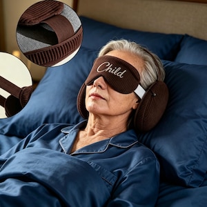 May include: A brown sleep mask with integrated ear muffs featuring the word Child embroidered in white script. The mask includes a wide elastic strap and a soft interior lining, designed to cover the eyes and ears for light and sound reduction.