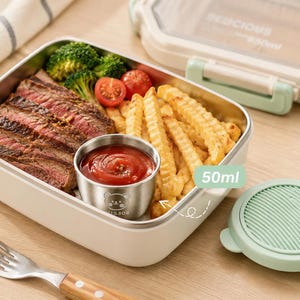 Puede incluir: Una fiambrera blanca y de acero inoxidable llena de bistec, brócoli, tomates cherry y patatas fritas onduladas. Un pequeño recipiente de acero inoxidable contiene ketchup. La tapa está parcialmente abierta y el texto "DELICIOUS" es visible.
