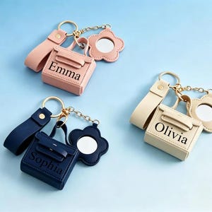 Könnte beinhalten: Drei personalisierte Schlüsselanhänger mit einem kleinen Handtaschen-Design, einem blütenförmigen Spiegel und einer Handschlaufe. Einer ist rosa mit "Emma", ein anderer ist marineblau mit "Sophia" und der dritte ist cremefarben mit "Olivia". Alle haben goldfarbene Hardware.