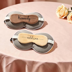 K&ouml;nnte beinhalten: Zwei personalisierte Schlafmasken mit den Namen "Kennedy" und "addison" aufgedruckt. Die Masken haben eine graue Basis, eine beige Mitte und wei&szlig;e elastische B&auml;nder. Sie liegen auf einer rosa Oberfl&auml;che, neben zwei gelben Rosen.