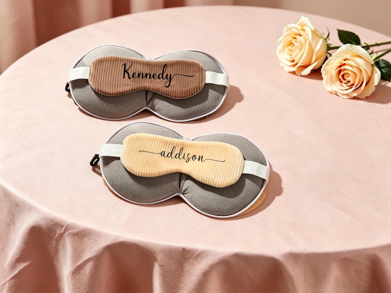 K&ouml;nnte beinhalten: Zwei personalisierte Schlafmasken mit den Namen "Kennedy" und "addison" aufgedruckt. Die Masken haben eine graue Basis, eine beige Mitte und wei&szlig;e elastische B&auml;nder. Sie liegen auf einer rosa Oberfl&auml;che, neben zwei gelben Rosen.