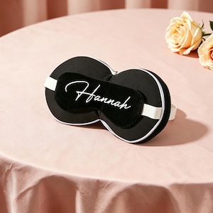 K&ouml;nnte beinhalten: Schwarze Schlafmaske mit wei&szlig;er Umrandung und wei&szlig;em Gummiband, personalisiert mit dem Namen "Hannah" in wei&szlig;er Schrift. Die Maske liegt auf einer rosa Oberfl&auml;che, mit zwei pfirsichfarbenen Rosen.