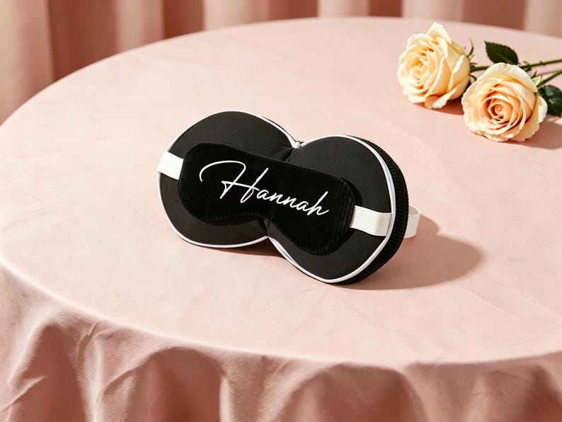 K&ouml;nnte beinhalten: Schwarze Schlafmaske mit wei&szlig;er Umrandung und wei&szlig;em Gummiband, personalisiert mit dem Namen "Hannah" in wei&szlig;er Schrift. Die Maske liegt auf einer rosa Oberfl&auml;che, mit zwei pfirsichfarbenen Rosen.