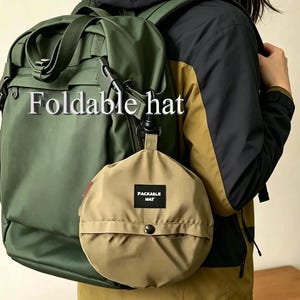 May include: A round, tan, foldable hat in a fabric pouch with a black label reading "PACKABLE HAT" attached to a large olive green backpack. The text "Foldable hat" is visible in the upper portion of the image, near the backpack straps.