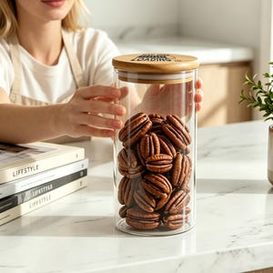 May include: A clear cylindrical glass storage jar filled with brown pecan halves, topped with a round bamboo lid featuring the black text "BABY LOADING..." printed on the surface. The container has a smooth, transparent finish.