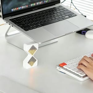 May include: A silver laptop sits on a silver laptop stand, with a white and gold hourglass-shaped device in front. A white keyboard with orange keys is in the foreground. The setup is on a white desk.
