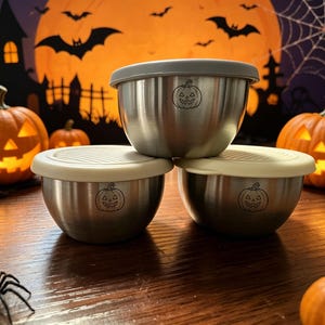 May include: Three stainless steel bowls with lids, each featuring a small engraved jack-o'-lantern pumpkin design on the front. The bowls are arranged in a stack with one on top and two below, showcasing a metallic finish and neutral-coloured lids.
