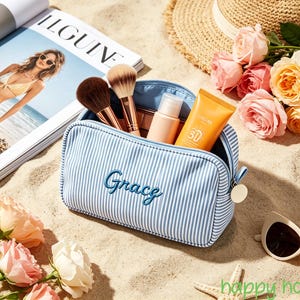 May include: A light blue and white striped zipped cosmetic bag with the name Grace embroidered in blue script on the front. The bag is open and contains two makeup brushes, a small beige bottle, and an orange sunscreen tube with 30 printed on it.