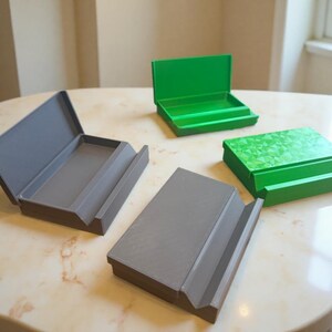Puede incluir: Cuatro pequeñas cajas rectangulares en tonos grises y verdes están dispuestas sobre una mesa con diseño de mármol. Una caja gris está abierta, revelando una bandeja interior. Dos cajas verdes están cerradas y una está abierta. Las cajas tienen un diseño simple y moderno.
