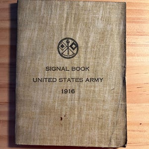 May include: A vintage Signal Book from the United States Army, dated 1916. The cover is tan and features a circular emblem with crossed flags. Text on the cover reads "SIGNAL BOOK UNITED STATES ARMY 1916". The book shows signs of wear.