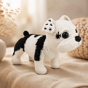 May include: A handmade crochet dog figurine in white and black. The dog has black spots on its ears and body, with large black button eyes and a black nose. The dog is standing on a neutral-colored surface.