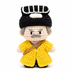 May include: A crocheted doll featuring a light-skinned face with a gray mustache, pink cheeks, and a stern expression. The doll wears a black hat with white vertical stripes and a bright yellow coat with a brown diagonal strap across the chest.