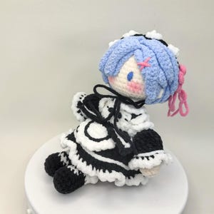 Puede incluir: Muñeca de ganchillo con un personaje de pelo azul, vestida con un traje de doncella blanco y negro. La muñeca tiene detalles rosas en el pelo y en las mejillas. La muñeca está sentada sobre una superficie blanca.