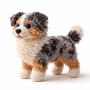 May include: A handmade plush dog toy in a standing position. The toy features a mix of white, gray, brown, and black yarn. The dog has a black nose, brown eyes, and a pink tongue. The tail is a fluffy pom-pom.