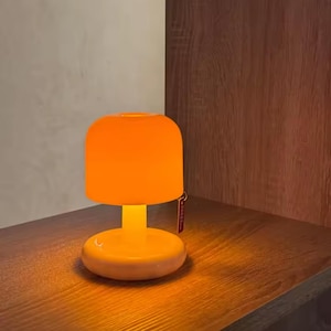Pode incluir: Uma pequena luminária de mesa laranja com uma cúpula arredondada e uma base circular. A luminária é feita de vidro e fica em uma superfície de madeira. Uma pequena etiqueta pende da luminária. A luminária emite um brilho laranja quente.