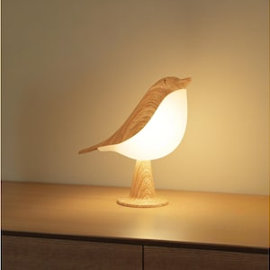 May include: A wooden bird-shaped table lamp with a warm, glowing light. The lamp features a light-colored wooden body and a white, egg-shaped light source. The lamp is on a wooden surface.