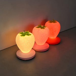 Puede incluir: Tres lámparas con forma de fresa con superficie texturizada y detalles de hojas verdes. Las lámparas están encendidas, con colores que van del blanco al naranja. Cada lámpara se asienta sobre una base redonda rosa, creando una exhibición de iluminación caprichosa.