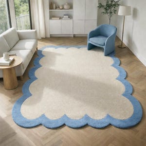Può includere: Un grande tappeto color crema con un bordo smerlato azzurro. Il tappeto è posizionato su un pavimento in legno in un soggiorno, con un divano bianco, una sedia blu e un tavolino visibili sullo sfondo. Il design del tappeto aggiunge un elemento decorativo morbido alla stanza.