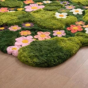 May include: A colourful rug depicting a garden scene with green, textured grass-like areas and embroidered flowers. The flowers have yellow centres and petals in shades of pink, orange, and white. The rug is placed on a light brown wooden floor.
