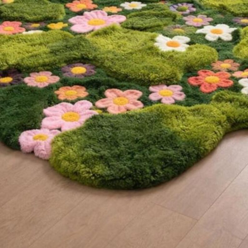May include: A colourful rug depicting a garden scene with green, textured grass-like areas and embroidered flowers. The flowers have yellow centres and petals in shades of pink, orange, and white. The rug is placed on a light brown wooden floor.