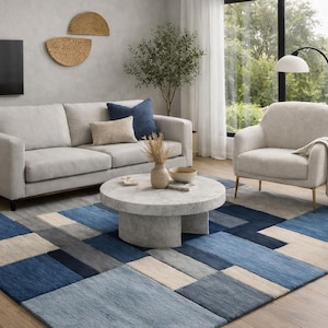 May include: A contemporary living room featuring a light grey sofa, armchair, and a circular marble coffee table. A blue and grey geometric rug is on the floor. Decorative items include cushions, vases, and wall art.