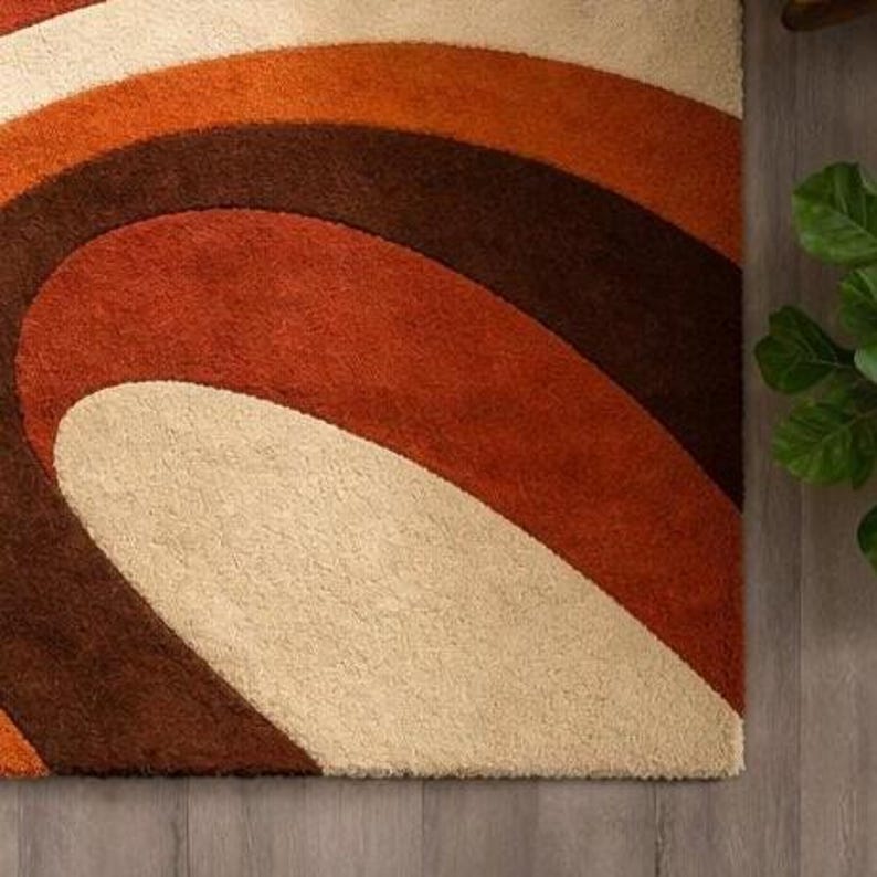 May include: A close-up of a retro-style rug with a swirling pattern. The rug features a cream, orange, brown, and rust color scheme. The design is reminiscent of the 1970s, with its bold, curved lines and warm color palette. The rug is made of a soft, textured material.