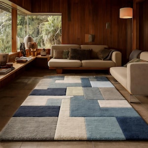 May include: A rectangular rug with a geometric pattern in shades of blue, grey, and cream. The rug is in a living room with wooden walls and a beige sofa. The rug's design features large, irregular blocks of colour.
