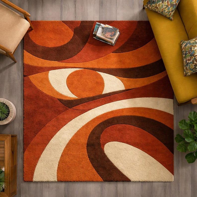 May include: A retro-style area rug with an abstract design in shades of orange, brown, and cream. The rug features curved lines and circular shapes, creating a bold, eye-catching pattern. The rug is rectangular and appears to be made of a soft, textured material.