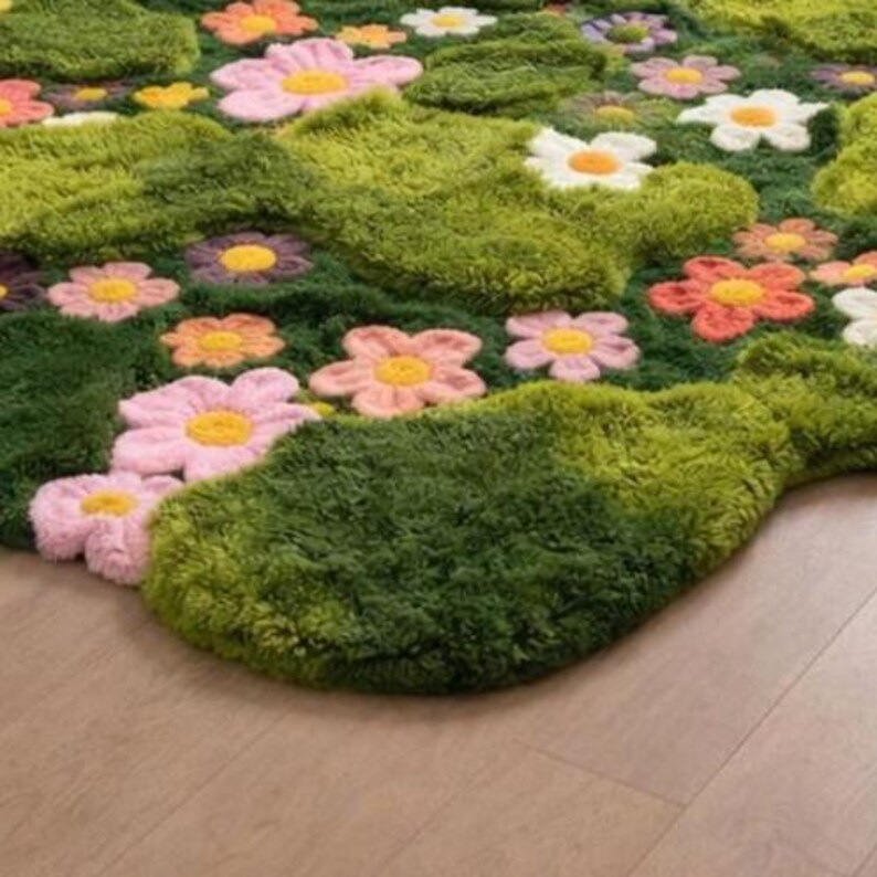 May include: A colourful, tufted rug with a meadow design. The rug has a green base with large, textured flower shapes in pink, orange, white, and purple, each with a yellow centre. The rug is placed on a light brown wooden floor.