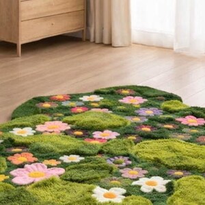 May include: A green, flower-shaped rug with a textured, grassy appearance. The rug features colourful, three-dimensional flower appliqu&eacute;s in shades of pink, yellow, orange, and white. The rug is placed on a wooden floor, with a wooden dresser in the background.