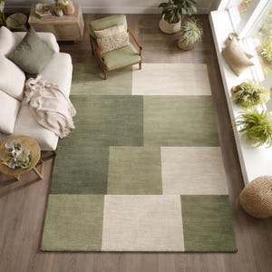 May include: An overhead shot of a geometric rug in shades of green and cream. The rug has a patchwork design with rectangular blocks of varying tones, creating a modern, textured look. It's placed on a wooden floor, with furniture visible in the background.