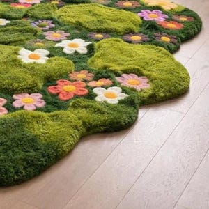 May include: A colourful, floral rug with a plush, green, moss-like texture. The rug features embroidered flowers in pink, orange, yellow, and white. The rug's irregular shape adds to its unique, artistic design. The rug is on a wooden floor.