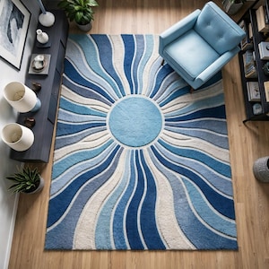 May include: A blue area rug with a sunburst design. The rug features concentric waves of varying shades of blue, from deep navy to light sky blue, radiating from a central light blue circle. The rug is placed on a wooden floor.