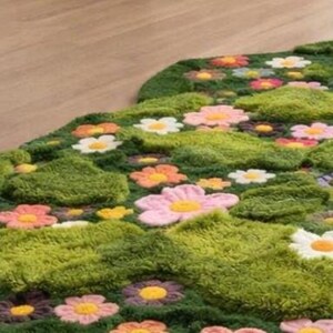 May include: A green, floral-shaped rug with a textured, grassy appearance. The rug features colourful, three-dimensional flower appliqu&eacute;s in shades of pink, orange, yellow, and white, creating a playful, garden-like design. The rug is placed on a light-coloured wooden floor.