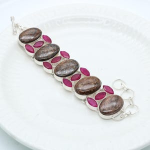 May include: A silver bracelet featuring oval-shaped brown stones and smaller, marquise-cut, deep pink stones. The bracelet is set on a white plate. The clasp is a toggle style.