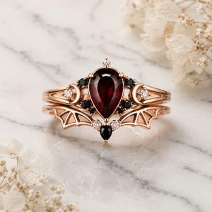 Puede incluir: Un anillo de oro rosa con un granate rojo oscuro en forma de pera, acentuado por pequeñas piedras negras y diamantes redondos transparentes. El diseño incorpora formas de media luna y motivos de alas de murciélago en capas.