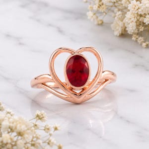 May include: A rose gold ring featuring a central oval-cut deep red gemstone set within a heart-shaped frame. The band is smooth and polished, with the heart design extending upwards to cradle the stone, creating a continuous, flowing metal silhouette.